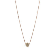 A delicate rose gold chain necklace featuring a small, raw, faceted diamond pendant, shown against a solid white background.