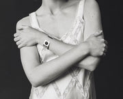 A black and white close-up of a person wearing a distinctive cuff bracelet with a circular center detail on their left wrist, paired with a lace-trimmed camisole against a dark background.