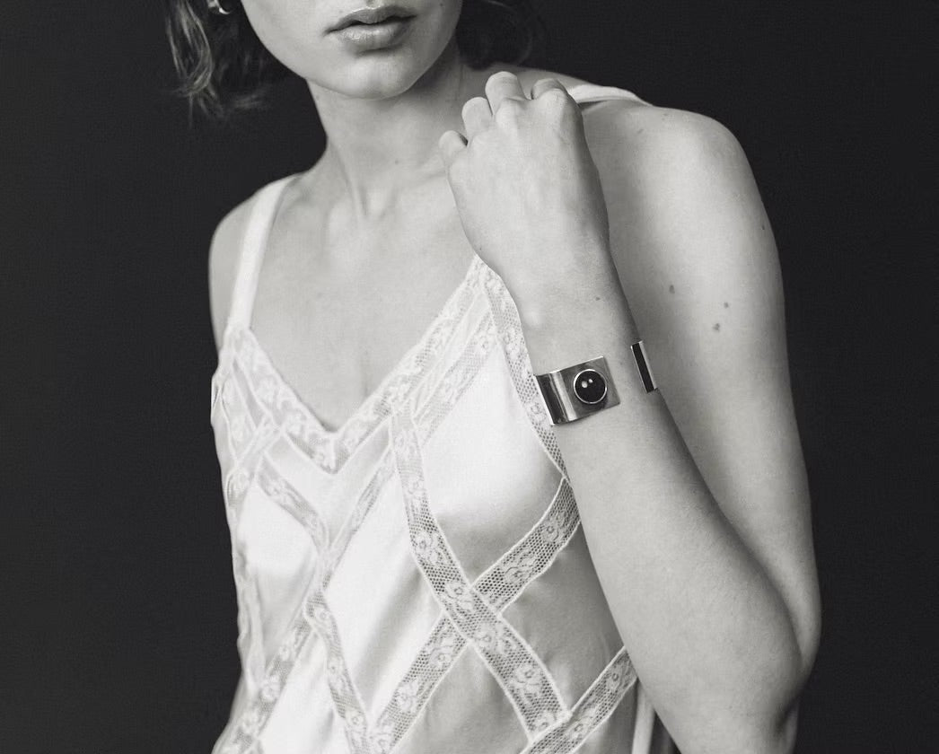 A black and white close-up of a woman wearing a metallic cuff bracelet with a circular face, paired with a lace-trimmed camisole against a dark background.