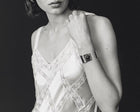 A black and white close-up of a woman wearing a metallic cuff bracelet with a circular face, paired with a lace-trimmed camisole against a dark background.