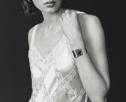 A black and white close-up of a woman wearing a metallic cuff bracelet with a circular face, paired with a lace-trimmed camisole against a dark background.