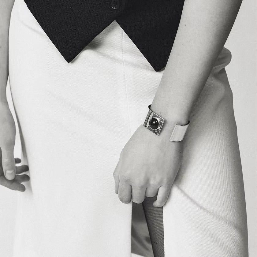 A close-up black and white photo of a person wearing a distinctive silver cuff bracelet with a black amber on their wrist, paired with a white skirt and dark top.