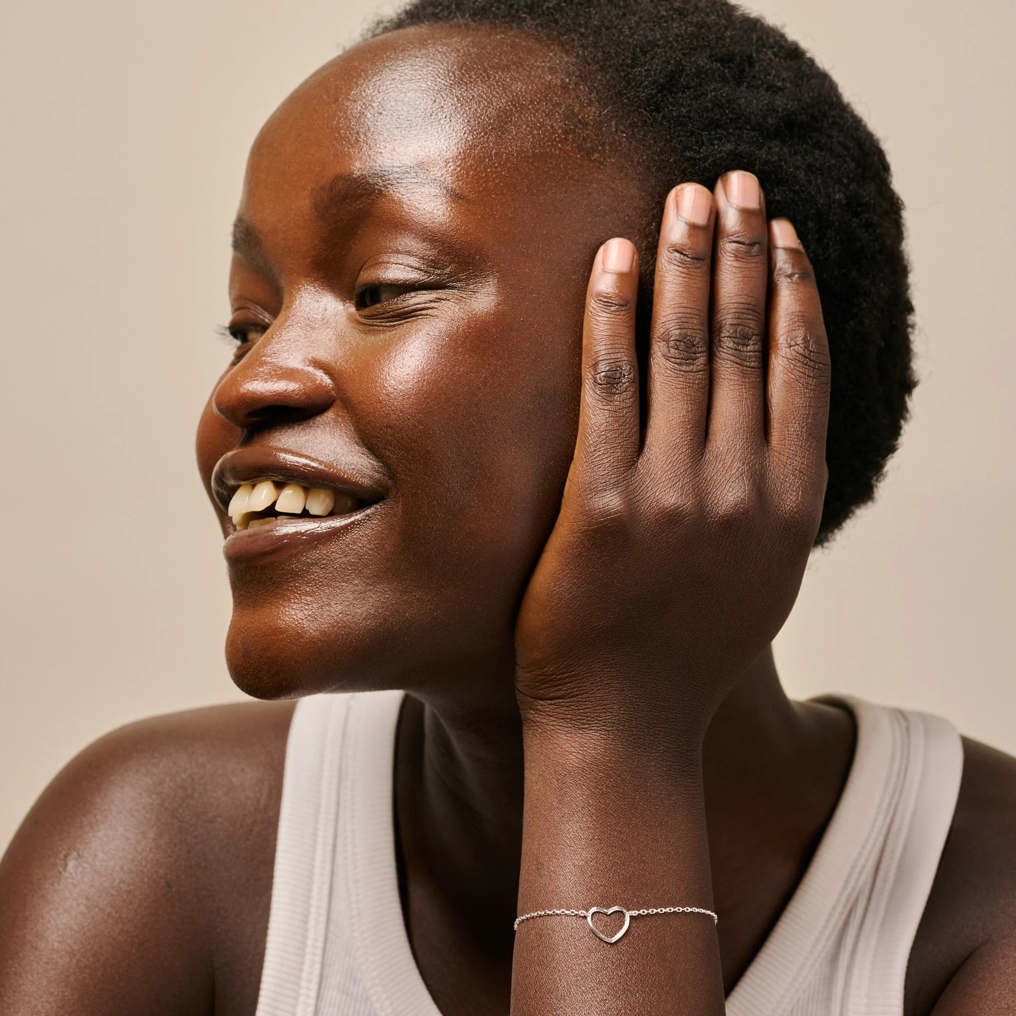 A close-up lifestyle shot of a smiling woman with short dark hair, wearing a delicate silver heart-link bracelet on her wrist, set against a warm beige background.