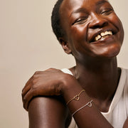 A close-up lifestyle shot of a smiling woman wearing two delicate heart-link bracelets, one gold and one silver, on her wrist, against a soft beige background.