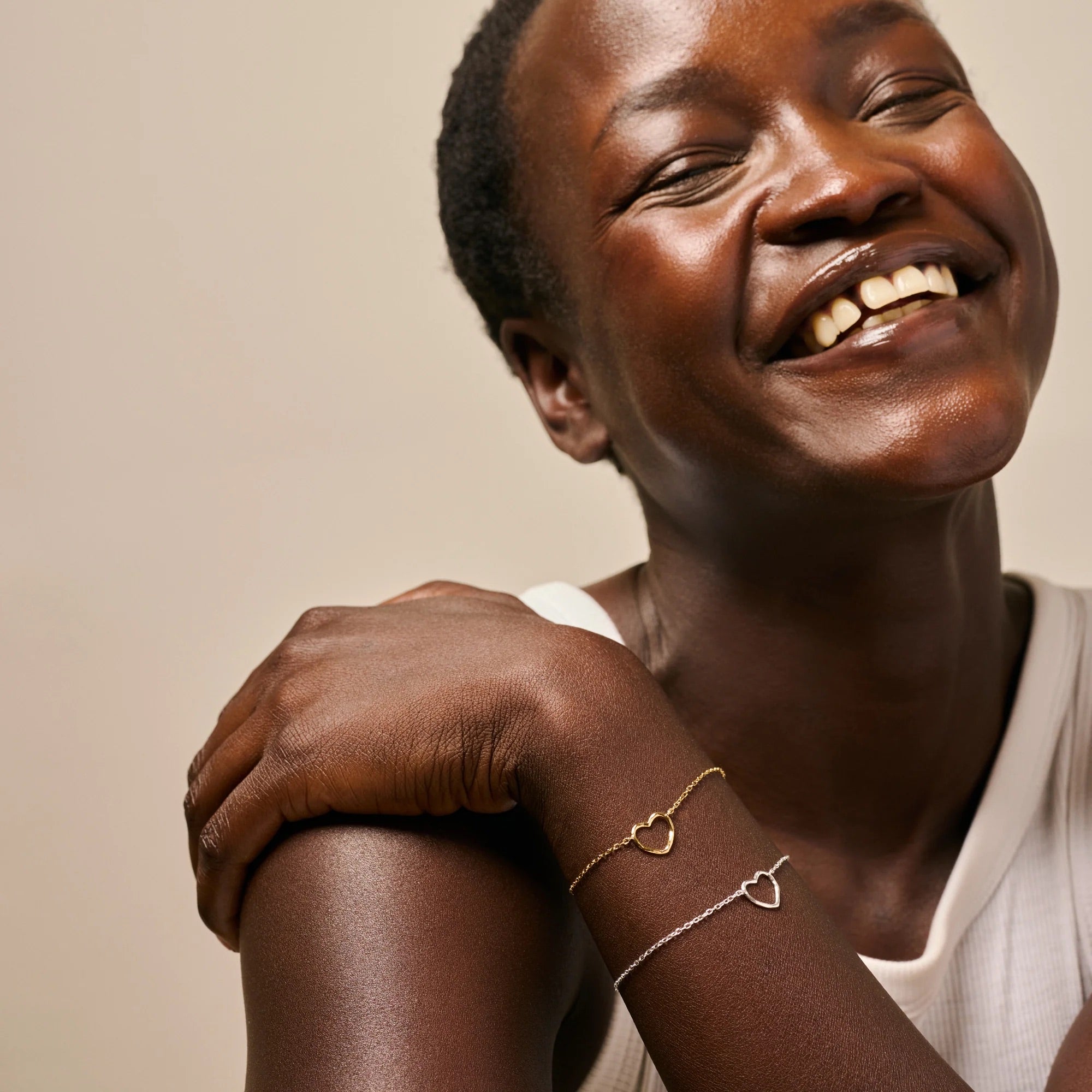 A close-up lifestyle shot of a smiling woman wearing two delicate heart-link bracelets, one gold and one silver, on her wrist, against a soft beige background.