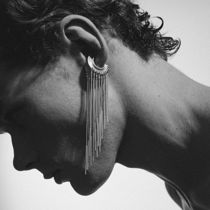 A close-up black and white portrait of a person in profile wearing a dramatic long dangling earring with multiple thin vertical strands and a circular hoop element.