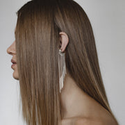 A woman shown in profile with long straight brown hair, wearing a dangling silver earring with multiple thin fringe strands against a plain light gray background.