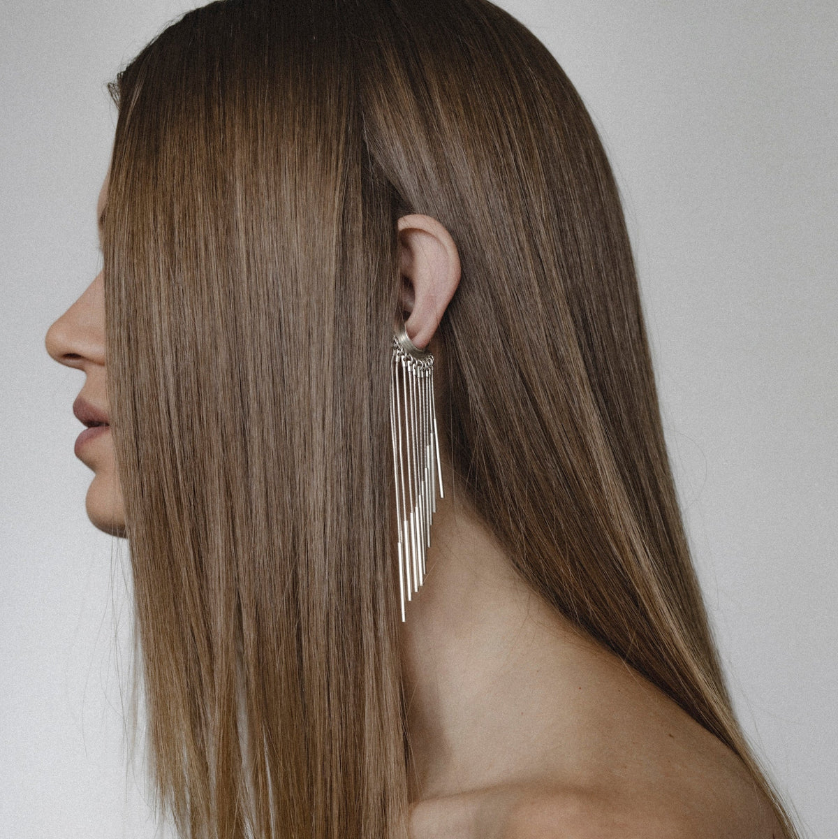 A woman shown in profile with long straight brown hair, wearing a dangling silver earring with multiple thin fringe strands against a plain light gray background.