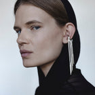 A close-up portrait of a model in profile wearing a black head covering and black top, with a long, dramatic silver dangle earring hanging from her ear against a plain light background.