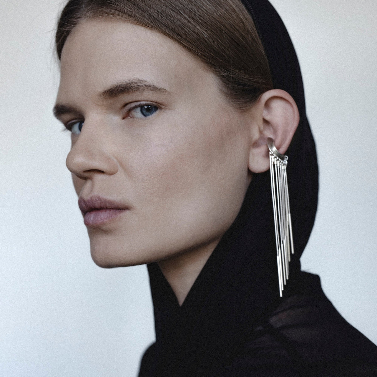 A close-up portrait of a model in profile wearing a black head covering and black top, with a long, dramatic silver dangle earring hanging from her ear against a plain light background.