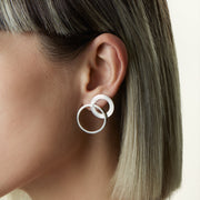 A close-up side profile of a model wearing a pair of silver double-ring hoop earrings against a clean, light beige background.