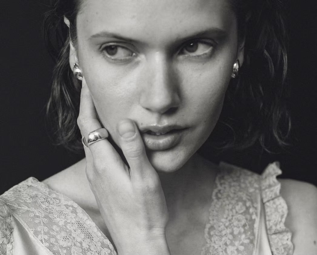 A black and white close-up portrait of a woman with her hand resting against her cheek, wearing a silver ring on her finger and small stud earrings, set against a dark background.