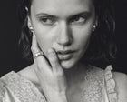 A black and white close-up portrait of a woman with her hand resting against her cheek, wearing a silver ring on her finger and small stud earrings, set against a dark background.