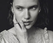 A black and white close-up portrait of a woman with her hand resting against her cheek, wearing a silver ring on her finger and small stud earrings, set against a dark background.