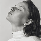 A black and white portrait of a woman with her eyes closed, wearing a white turtleneck and a small, round silver oval earring, set against a plain light gray background.