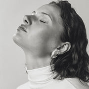 A black and white portrait of a woman with her eyes closed, wearing a white turtleneck and a small, round silver oval earring, set against a plain light gray background.