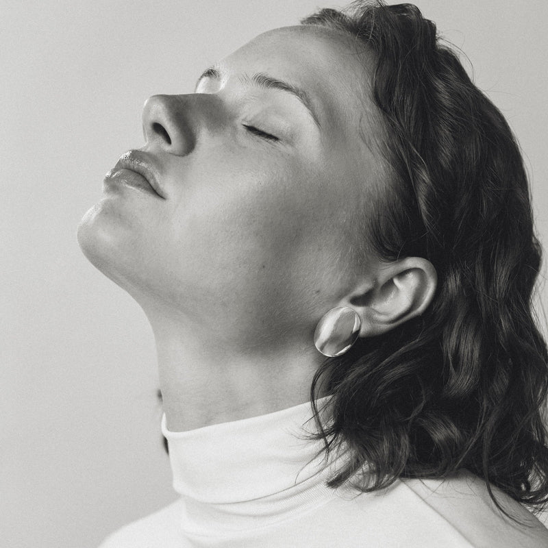 A black and white portrait of a woman with her eyes closed, wearing a white turtleneck and a small, round silver oval earring, set against a plain light gray background.