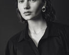 A black and white portrait of a woman wearing a black button-up shirt and a large circular black earring, set against a dark studio background.