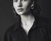 A black and white portrait of a woman wearing a black button-up shirt and a large circular black earring, set against a dark studio background.
