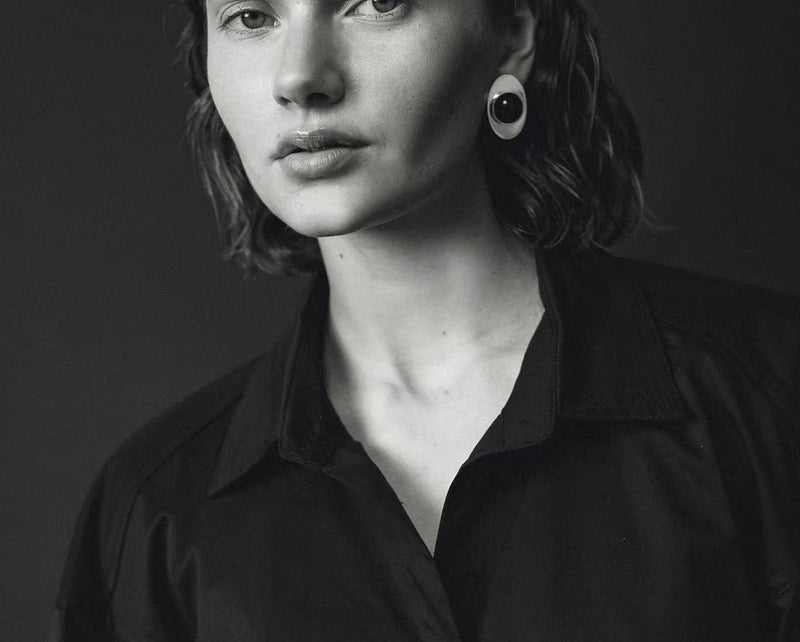 A black and white portrait of a woman wearing a black button-up shirt and a large circular black earring, set against a dark studio background.
