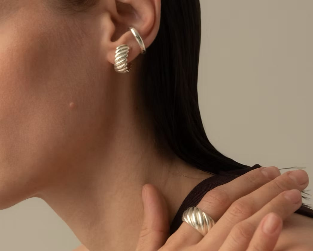 A close-up of a model wearing a silver twisted hoop earring in her ear and a matching silver twisted ring on her finger, set against a neutral beige background.