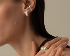A close-up of a model wearing a silver twisted hoop earring in her ear and a matching silver twisted ring on her finger, set against a neutral beige background.