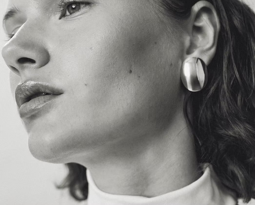 A close-up black and white photograph of a woman wearing a large, oval, polished silver earring.