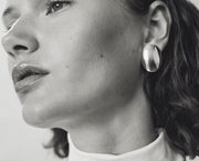 A close-up black and white photograph of a woman wearing a large, oval, polished silver earring.