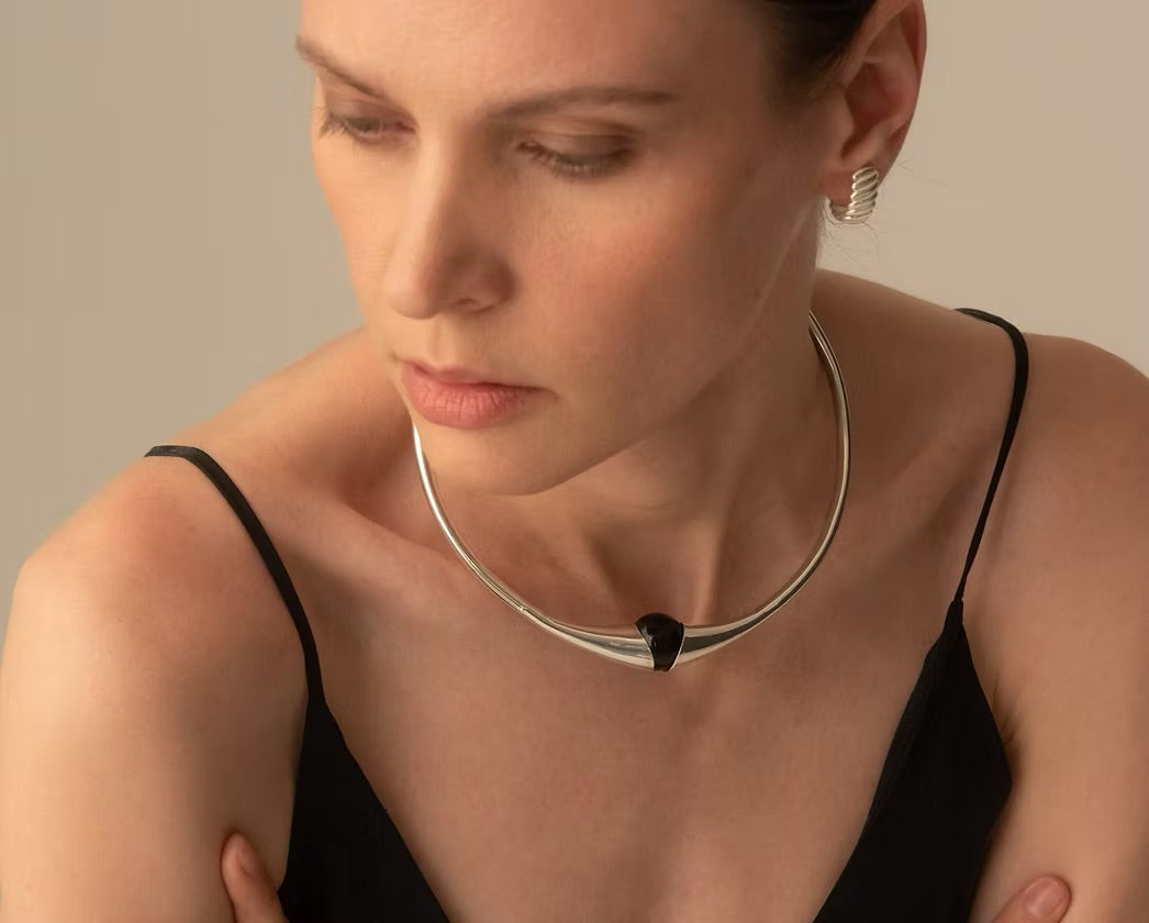 A close-up of a woman wearing a polished silver choker necklace with a small black triangular accent at the center, paired with a black camisole top against a warm beige background.
