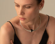 A close-up of a woman wearing a polished silver choker necklace with a small black triangular accent at the center, paired with a black camisole top against a warm beige background.