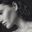 A close-up black and white portrait of a woman in profile, wearing a small hoop earring and a delicate chain necklace against a dark background.