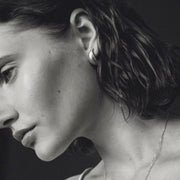 A close-up black and white portrait of a woman in profile, wearing a small hoop earring and a delicate chain necklace against a dark background.