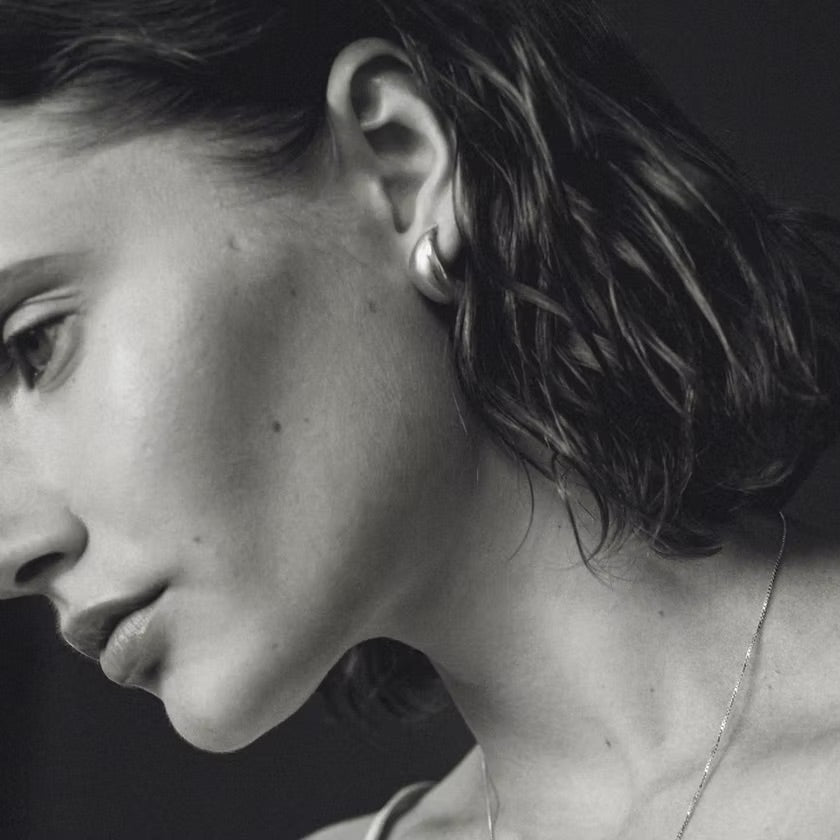 A close-up black and white portrait of a woman in profile, wearing a small hoop earring and a delicate chain necklace against a dark background.