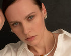 A close-up portrait of a woman wearing a chunky silver-toned metal collar necklace and matching textured hoop earrings, paired with a white collared blouse against a dark background.