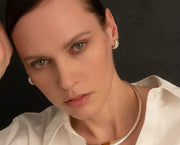 A close-up portrait of a woman wearing a chunky silver-toned metal collar necklace and matching textured hoop earrings, paired with a white collared blouse against a dark background.