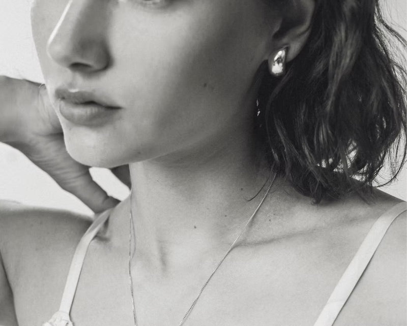 A close-up black and white photograph of a woman wearing a delicate silver-toned necklace and a small round stud earring.
