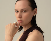 A close-up portrait of a woman wearing a silver huggie hoop earring with a textured, layered design. She is wearing a dark brown sleeveless top against a solid, warm beige background.