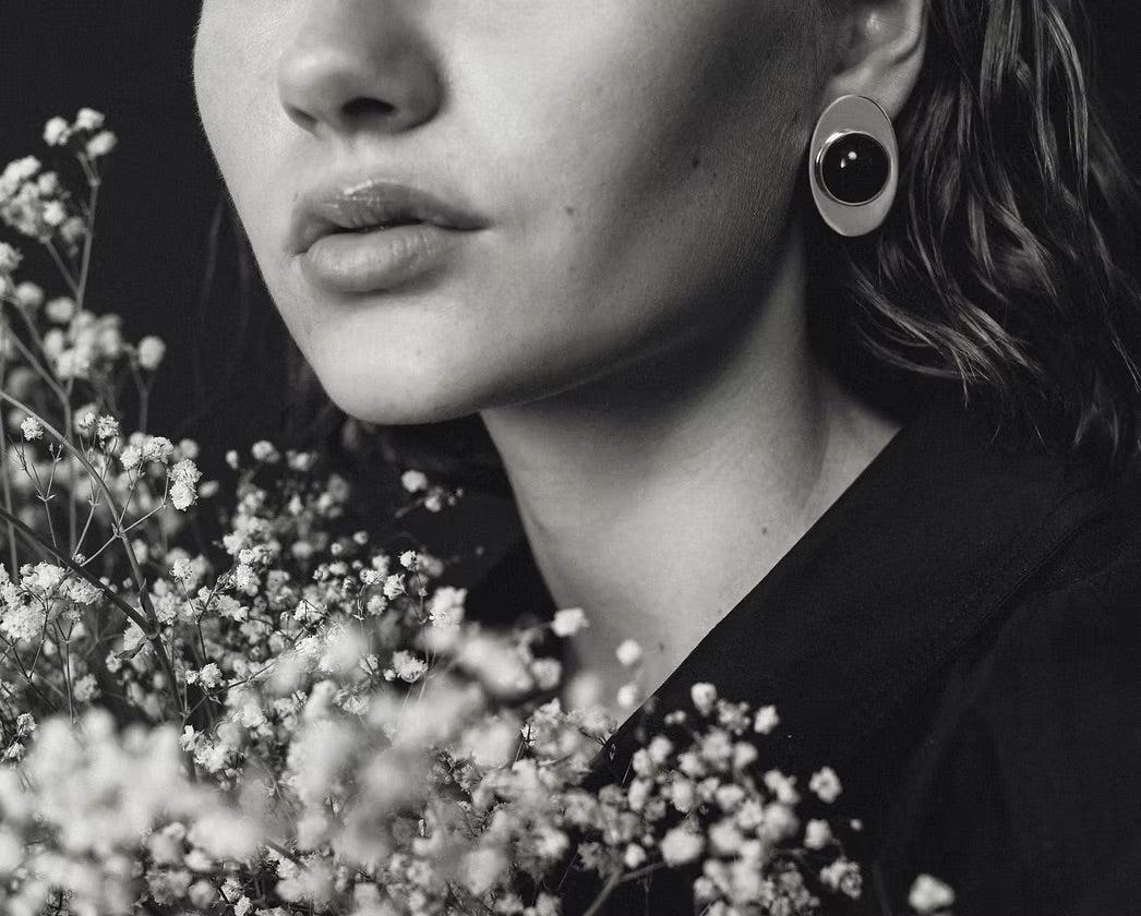A close-up black and white photograph of a woman wearing a distinctive circular earring with a dark amber and light outer ring, styled with baby's breath flowers.