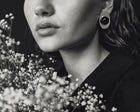 A close-up black and white photograph of a woman wearing a distinctive circular earring with a dark amber and light outer ring, styled with baby's breath flowers.