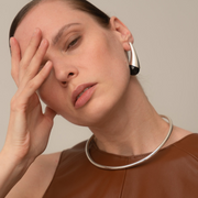 A close-up lifestyle shot of a woman wearing a chunky silver collar necklace and a matching silver hoop earring, posing with her hand on her face against a solid beige background.