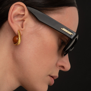 Close-up side profile of a woman wearing black sunglasses with gold-plated silver double-stripe detailing on the temple and a gold hoop earring with a brown bead.