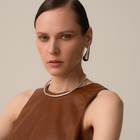 A close-up portrait of a model wearing a chunky silver-toned metal collar necklace and a matching silver teardrop earring, paired with a brown leather top against a neutral beige background.