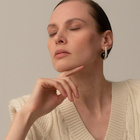 A close-up portrait of a woman wearing a small silver hoop earring with a black disc accent, paired with a cream cable-knit sweater against a neutral beige background.
