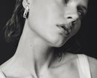 A close-up black and white photograph of a woman wearing a geometric silver earring, with her face and neck prominently shown against a dark background.