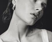 A close-up black and white photograph of a woman wearing a geometric silver earring, with her face and neck prominently shown against a dark background.