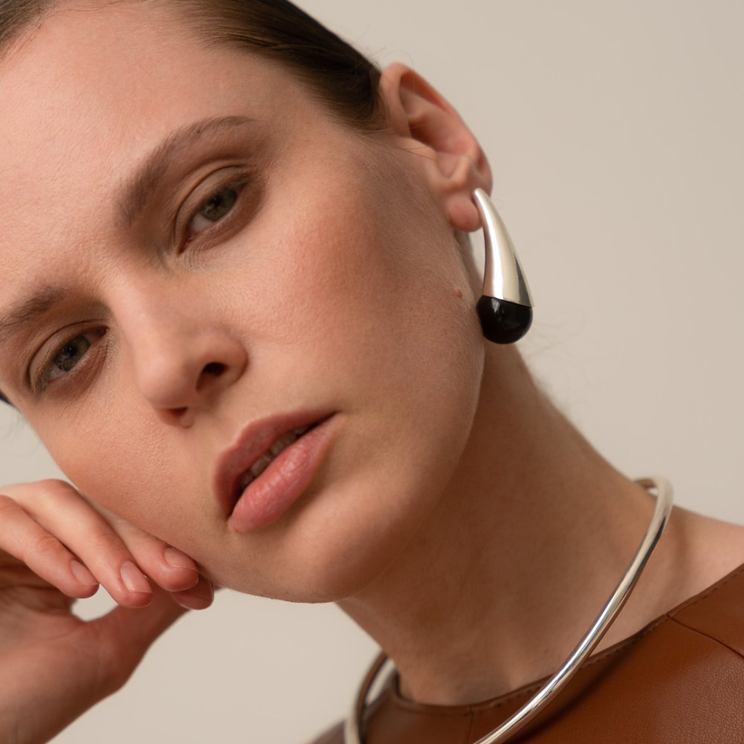Close-up of a woman wearing a sculptural silver earring with a black end, paired with a silver choker necklace and a brown top, against a neutral beige background.