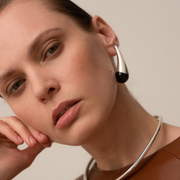 Close-up of a woman wearing a sculptural silver earring with a black end, paired with a silver choker necklace and a brown top, against a neutral beige background.