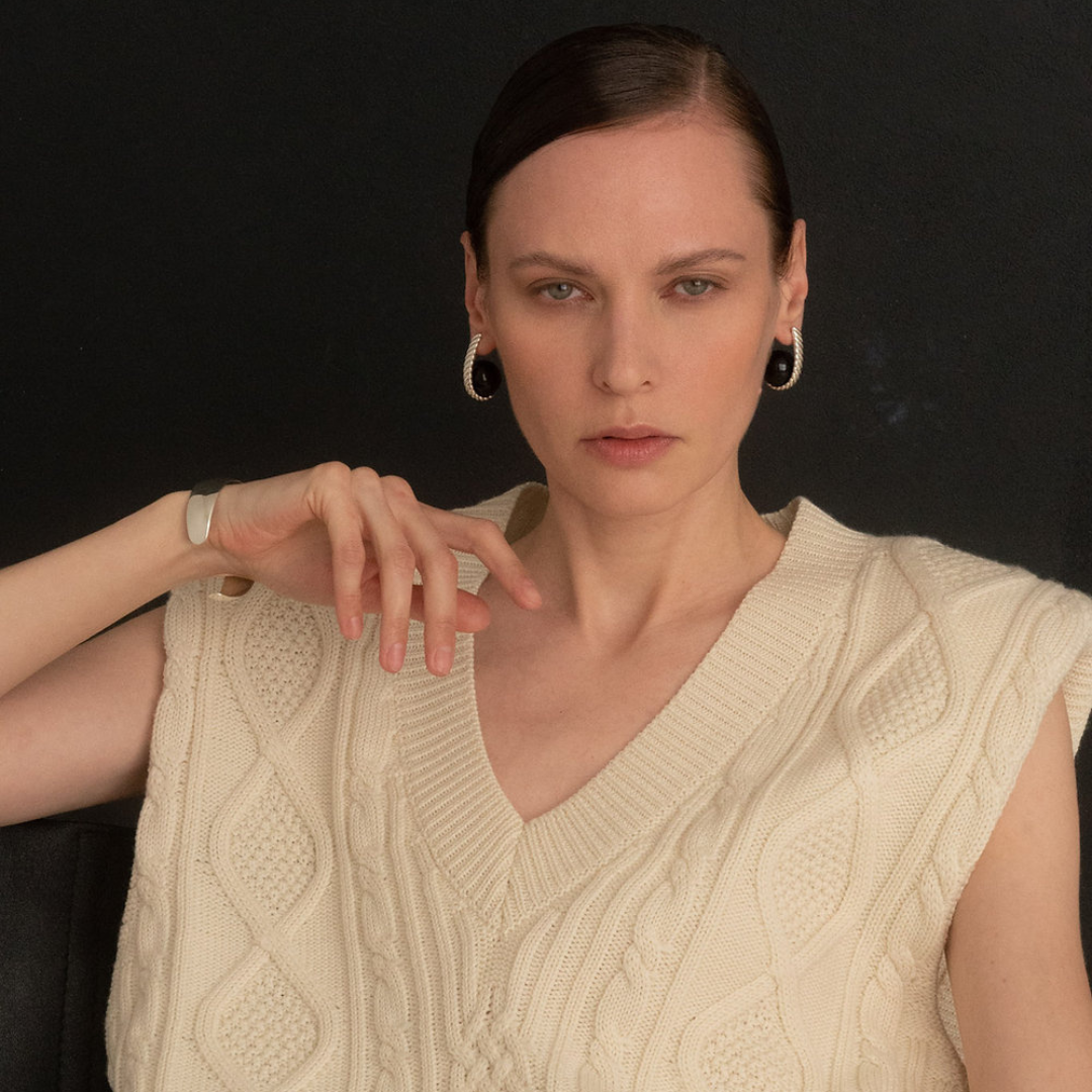 A woman with dark hair pulled back wears a cream cable-knit sleeveless V-neck sweater vest against a dark background. She is also wearing black hoop earrings and a silver-toned watch.