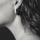 A close-up black and white photograph of a person's ear wearing a modern, curved earring with a dark circular amber and a silver-toned outer edge.