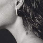 A close-up black and white photograph of a person's ear wearing a modern, curved earring with a dark circular amber and a silver-toned outer edge.